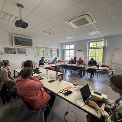 Teilnehmende der Cyberangriff-Krisenübung des Burgenlandkreises arbeiten konzentriert an Laptops in einem Besprechungsraum. Im Hintergrund stehen Mitarbeitende des Gesundheitsamts und des Amtes für Bevölkerungsschutz vor einer großen Leinwand und einer Einsatzkarte.