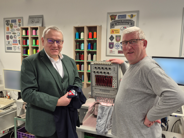 Landrat Götz Ulrich steht mit Ralf Single vor einer Stickmaschine in der Werkstatt der Stickerei Single in Dietrichsroda. Im Hintergrund bunte Garnrollen und Muster.