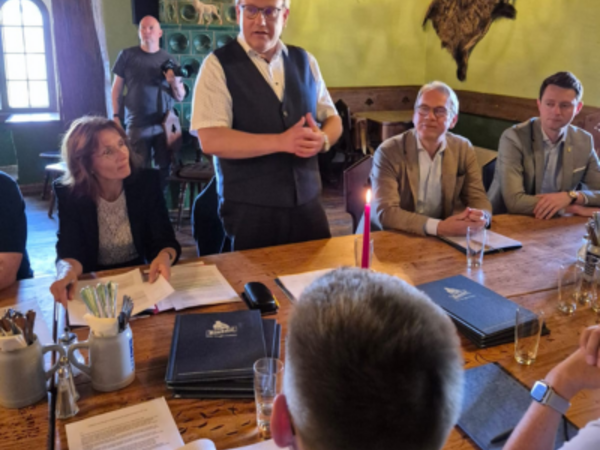 Landrat Götz Ulrich steht in einem historischen Raum der Rudelsburg und spricht zu einer Gruppe von Personen am Tisch, darunter Sachsen-Anhalts Innenministerin Dr. Tamara Zieschang, Thüringens Innenminister Georg Maier und weitere Landräte. Im Raum: Jagdtrophäen an der Wand, grüne Kachelöfen und Tageslicht durch ein Rundbogenfenster.
