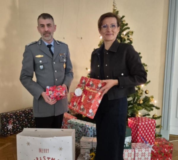 Oberstapotheker Martin Melcher und Dr. Ariane Körner stehen vor einem geschmückten Weihnachtsbaum mit Geschenken und halten Päckchen in den Händen.